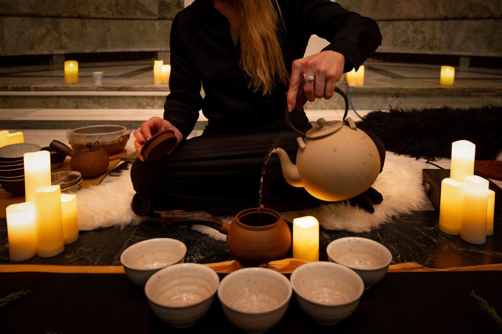 A seated person pours tea into tea bowls. Candles are lit in the area.